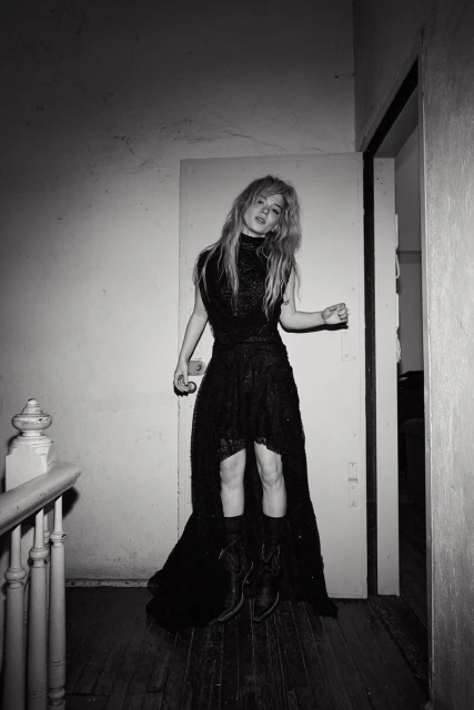 Young woman with blonde hair stands in dark dress and boots, posing in a hallway with an open doorway in a black and white photo.