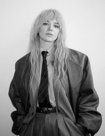 Black and white portrait of a woman with blonde hair, wearing an oversized suit jacket and tie, standing in front of a white wall.