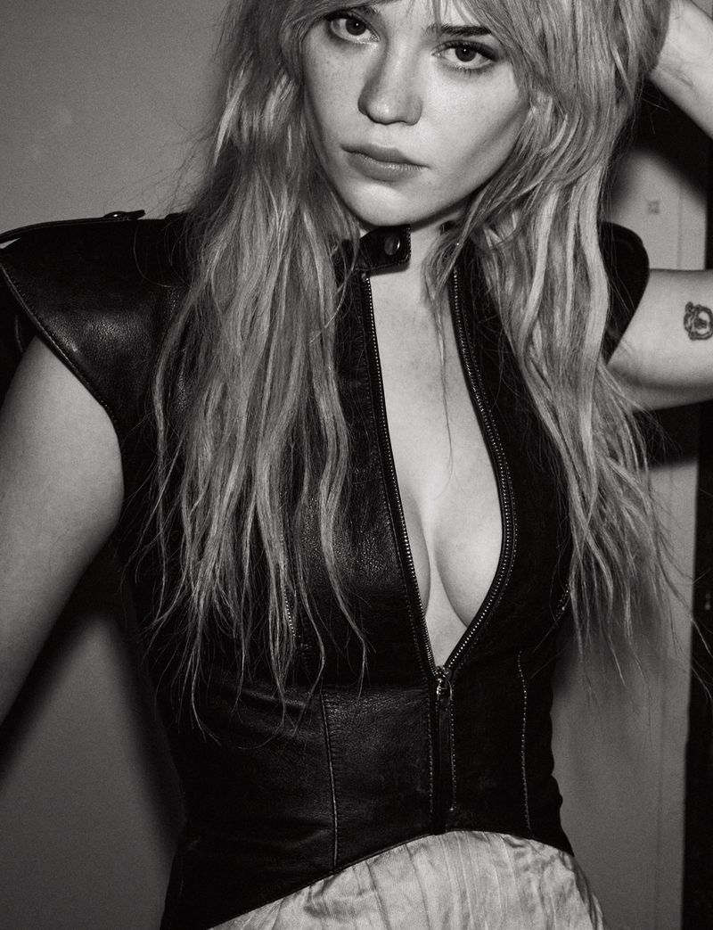 Black and white portrait of a fair-skinned woman with long blonde hair, wearing a leather vest and a neutral-toned skirt.