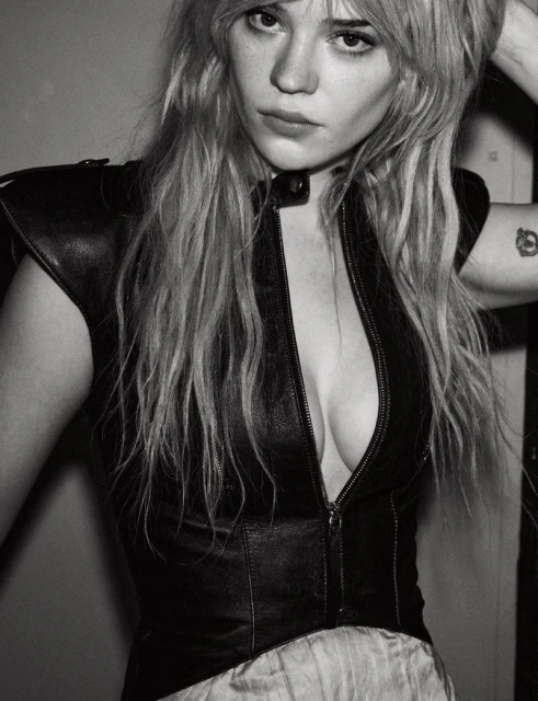 Black and white portrait of a fair-skinned woman with long blonde hair, wearing a leather vest and a neutral-toned skirt.