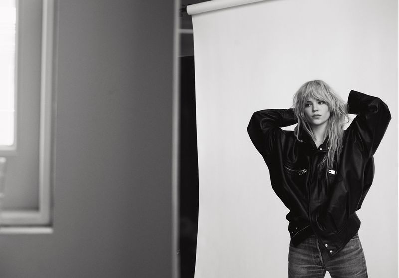 Monochrome portrait of a blonde woman in a black jacket, hands behind her head, against a white background in a photo studio.