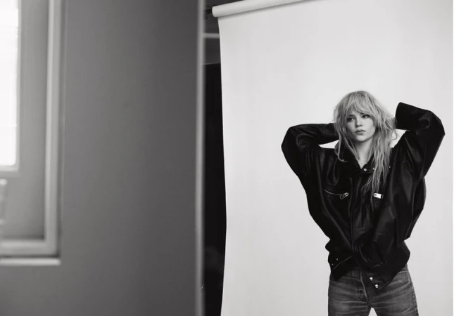 Monochrome portrait of a blonde woman in a black jacket, hands behind her head, against a white background in a photo studio.