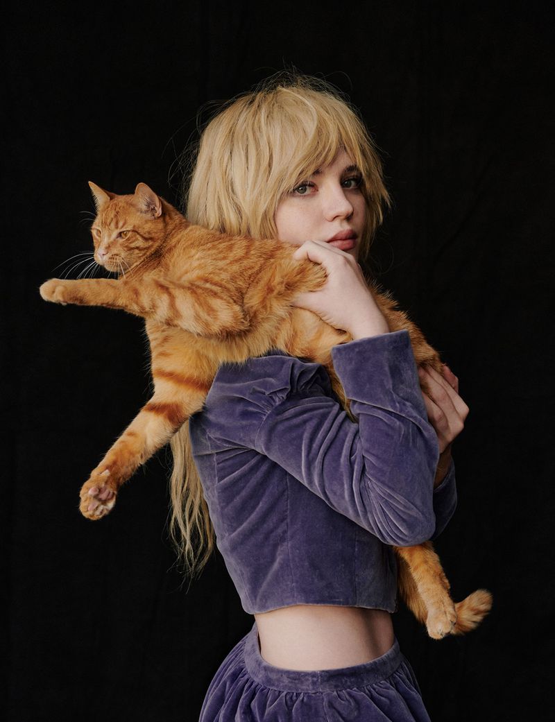 Portrait of a fair-skinned young woman with a fringe and long blond hair holding an orange tabby cat against a black backdrop.