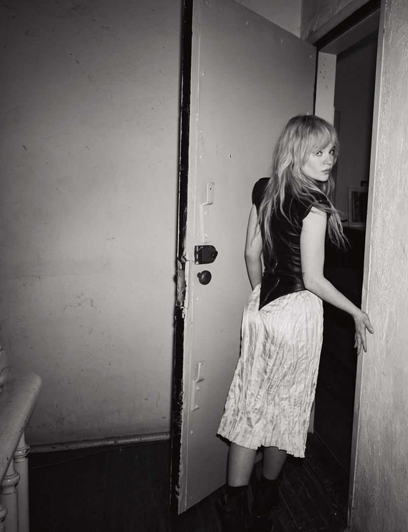 Black-and-white portrait of a woman in a black top and white skirt standing in a doorway, looking over her shoulder at the camera.
