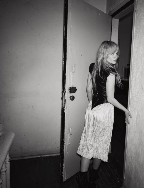 Black-and-white portrait of a woman in a black top and white skirt standing in a doorway, looking over her shoulder at the camera.