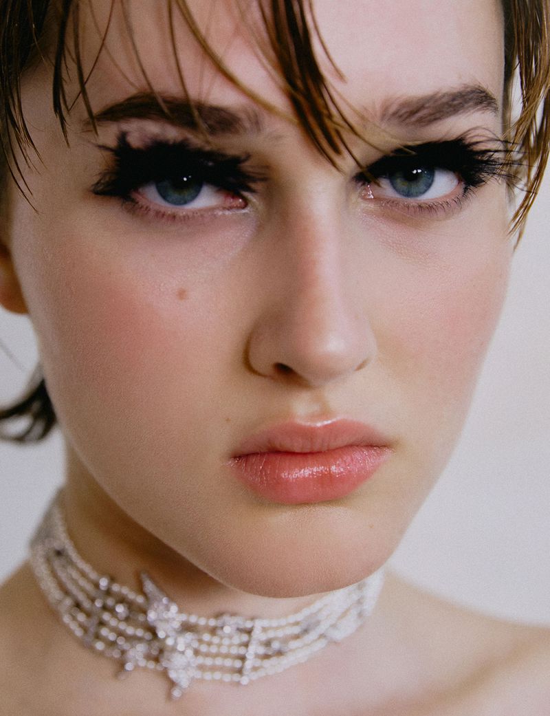 Close-up portrait of a woman with wet short hair and elaborate false eyelashes, wearing a multi-strand pearl and crystal choker.