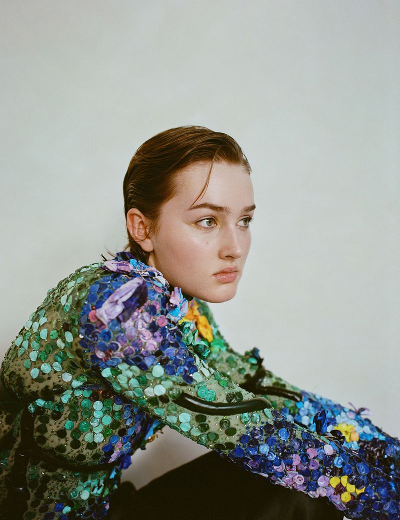 Portrait of a young woman with slicked-back hair wearing an embellished, jewel-toned blouse against a plain background in studio lighting.