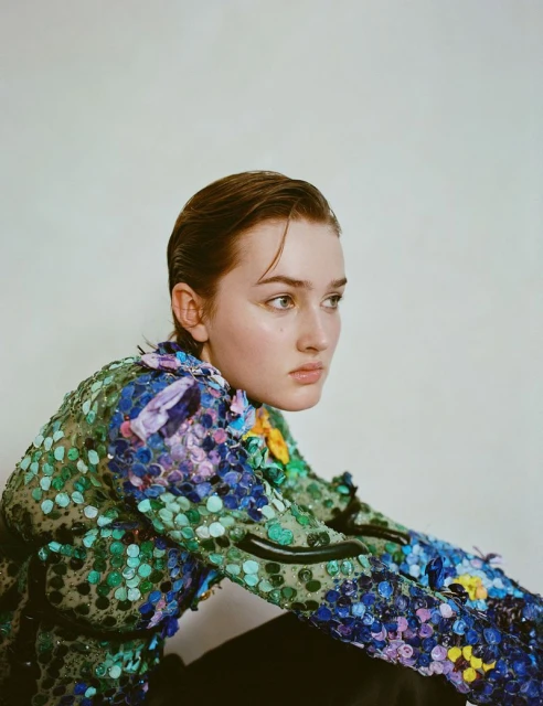 Portrait of a young woman with slicked-back hair wearing an embellished, jewel-toned blouse against a plain background in studio lighting.