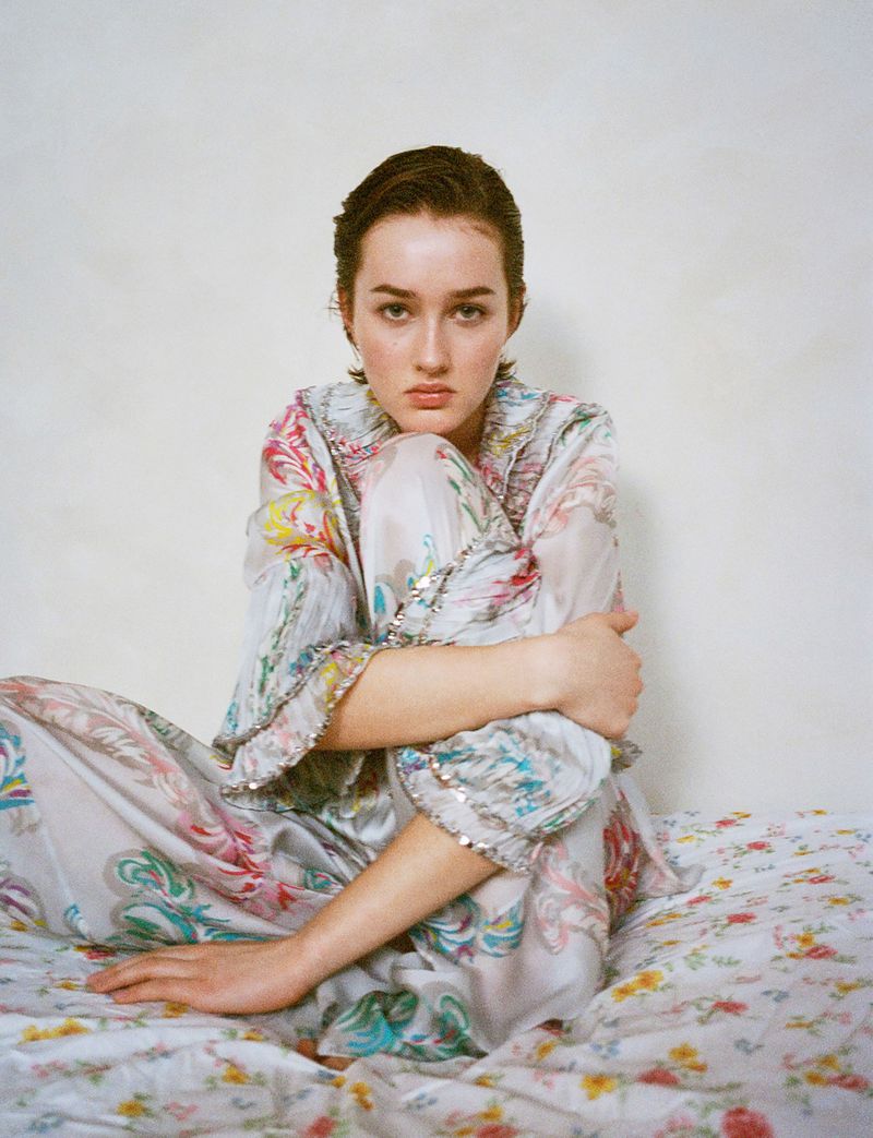Young woman in a floral patterned silk pajama set sits cross-legged on a matching bedspread in an indoor setting with soft, natural lighting.
