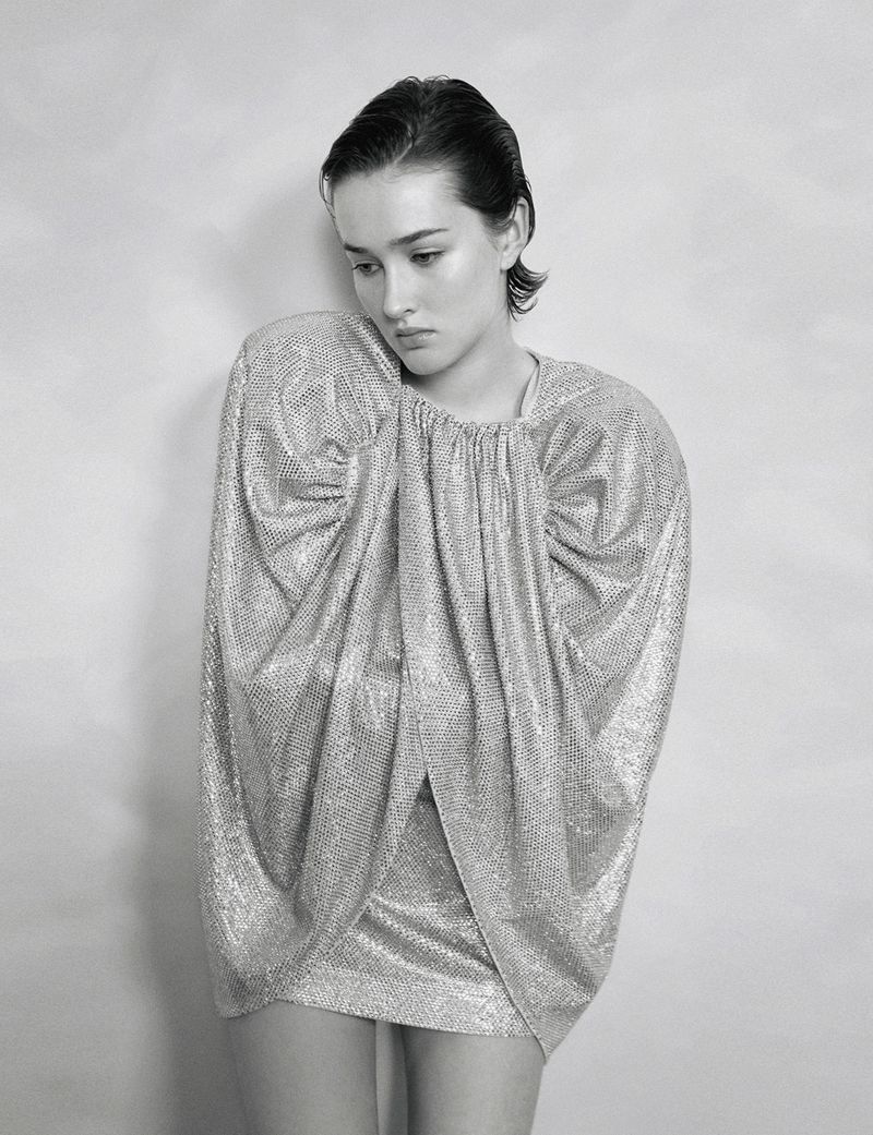 Monochrome fashion portrait of a model wearing a sparkly silver dress with gathered details against a neutral wall in a photo studio.