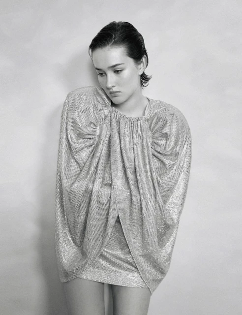 Monochrome fashion portrait of a model wearing a sparkly silver dress with gathered details against a neutral wall in a photo studio.