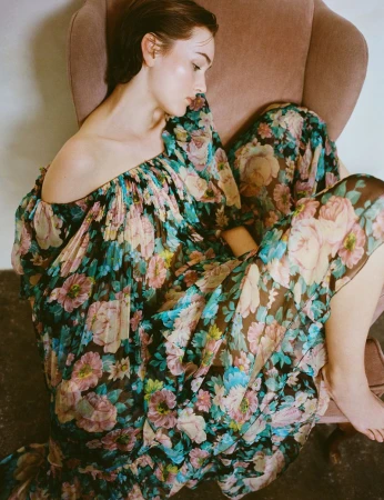 A young woman lounges in a pink armchair, wearing a flowing, floral-patterned off-the-shoulder dress in a brightly lit indoor setting.