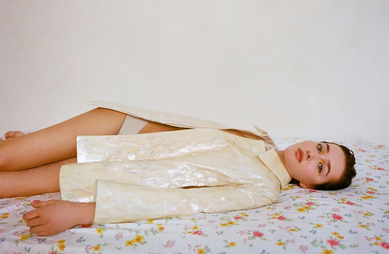 Adult female model poses lying on a floral patterned bed wearing an open iridescent jacket against a neutral white wall indoors.