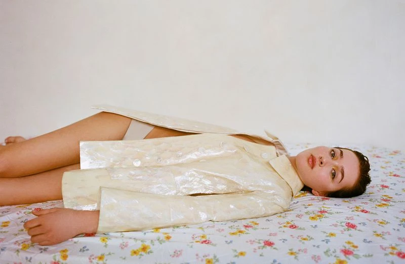 Adult female model poses lying on a floral patterned bed wearing an open iridescent jacket against a neutral white wall indoors.