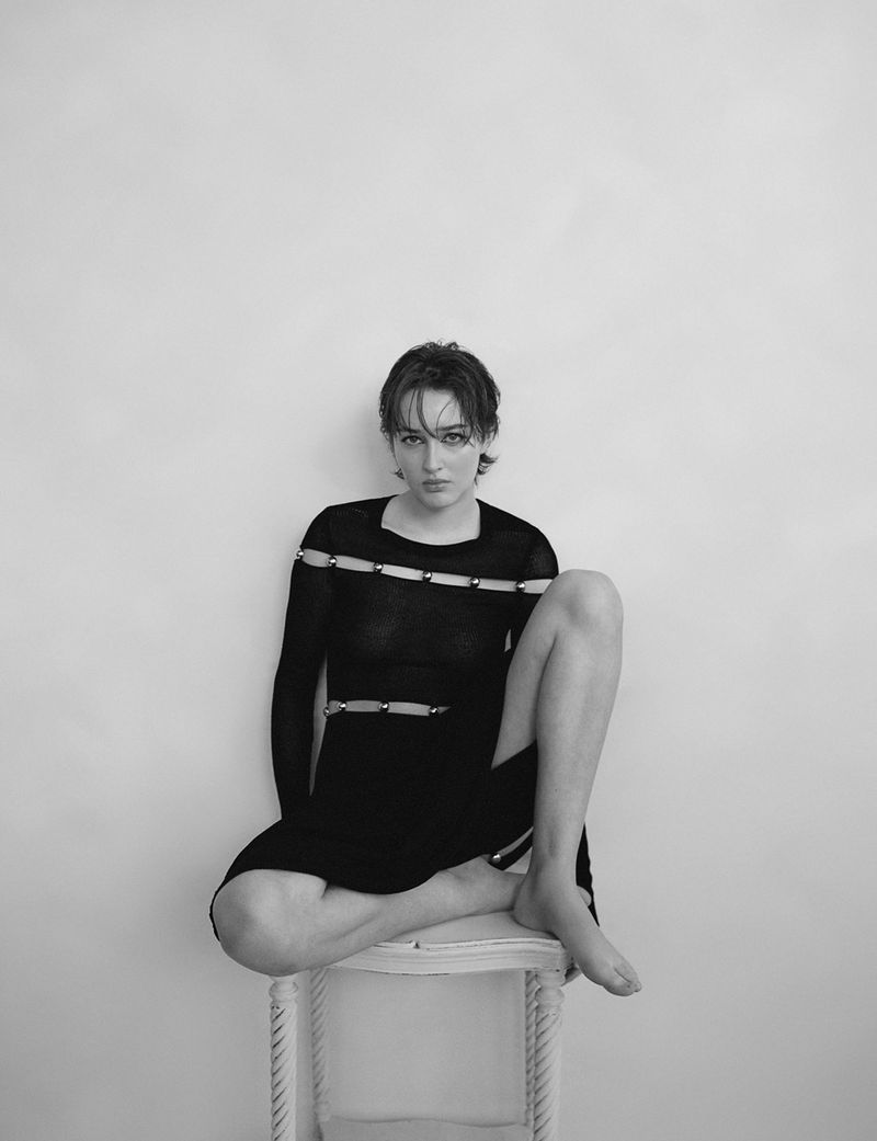 Model with short dark hair wearing a black dress seated on a white stool against a plain studio background.