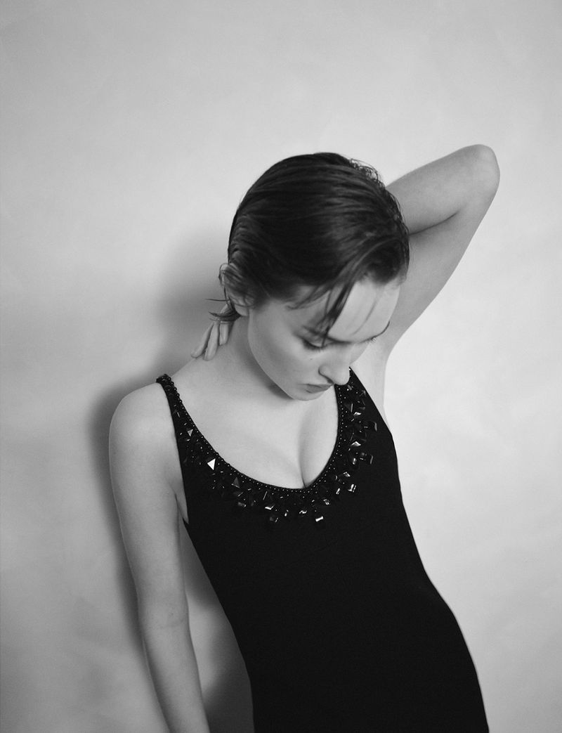 A young woman with dark hair and fair skin poses in a black sleeveless dress against a light grey backdrop, looking downwards.