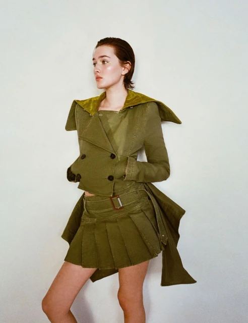 A slender young woman poses against a neutral backdrop, showcasing an olive-green ensemble featuring a short skirt and a cropped jacket.