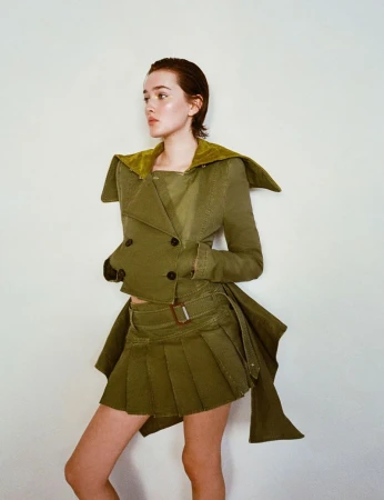 A slender young woman poses against a neutral backdrop, showcasing an olive-green ensemble featuring a short skirt and a cropped jacket.