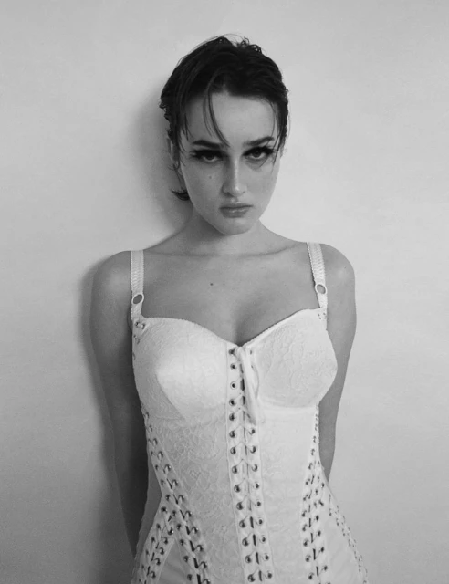 Black-and-white portrait of a young woman with dark hair, wearing a white corset dress against a neutral wall. Fashion photography.