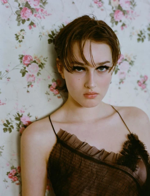 Portrait of a brunette woman with heavy makeup and a ruffled top, posing indoors against a floral wallpaper backdrop in a fashion editorial.
