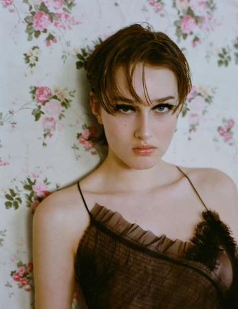 Portrait of a brunette woman with heavy makeup and a ruffled top, posing indoors against a floral wallpaper backdrop in a fashion editorial.