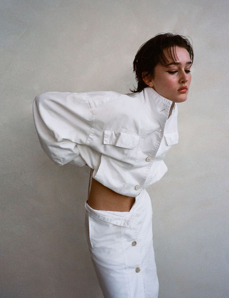A young woman poses in white separates against a light neutral background, eyes downcast, in a fashion-forward, minimalist style portrait.