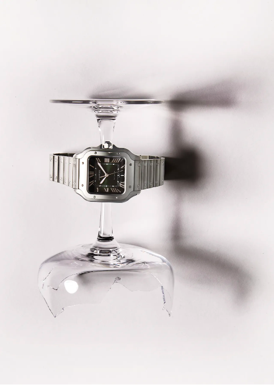 Silver wristwatch showcased between an intact wine glass top and a shattered glass base against a bright, shadow-casted backdrop.