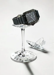 Black square wristwatch resting on the jagged stem of a broken crystal wine glass against a white background.