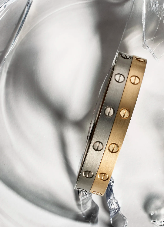 Two metal bracelets, one silver and one gold, with round screw-head details, on a clear reflective surface, jewelry product shot.