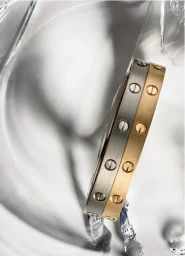 Two metallic bangles with screw motifs resting against transparent reflective glass surfaces in an editorial macro shot.