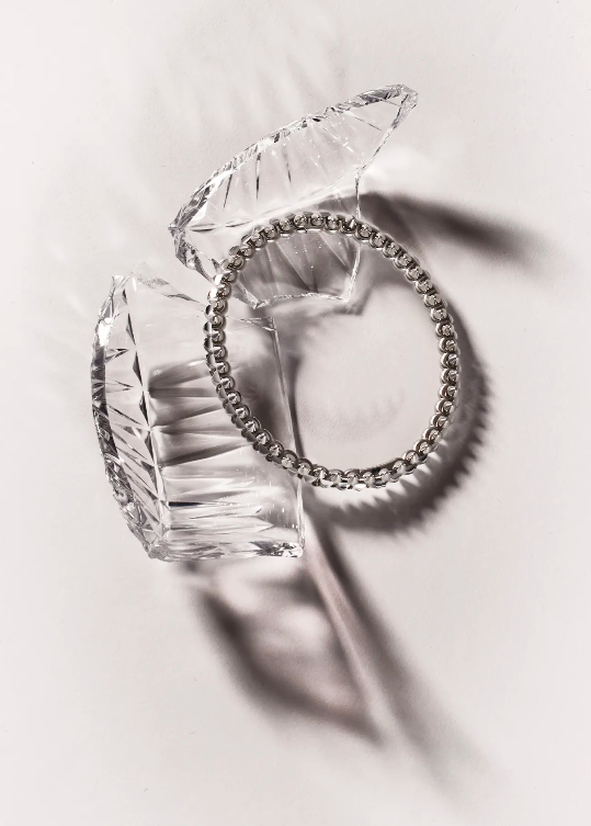 Elevated studio shot of a silver beaded bracelet resting inside an open crystal trinket box against a white backdrop with shadows.