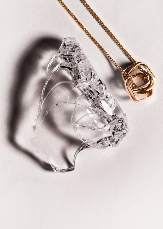 Still life featuring a clear shard of glass and a gold necklace with a three-ring pendant on a white surface, emphasizing texture and light.