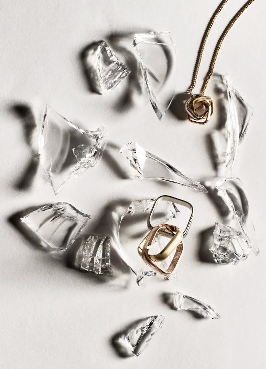 Jewelry still life features gold necklaces and rings scattered among shattered glass pieces on a bright white surface, creating a striking image.
