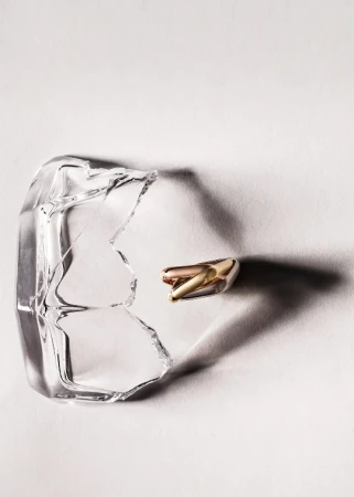 Close-up studio shot of a broken glass and two gold and white capsules, artfully arranged on a white surface with shadow play.