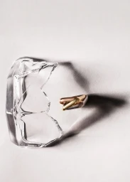 Close-up still life of a gold ring resting near a shattered piece of glass on a neutral grey textured background.