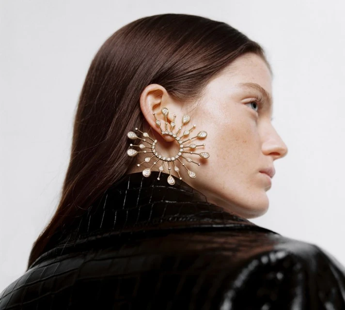Woman with dark hair wearing an elaborate gold sunburst earring with gems against a neutral background in a beauty fashion shot.
