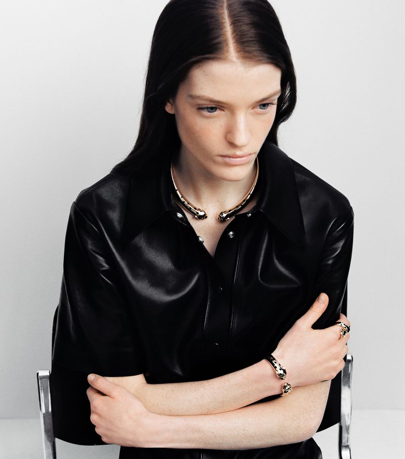 Model in black leather shirt wearing gold choker and bracelets against a white background