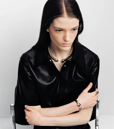 Model in black leather shirt wearing gold choker and bracelets against a white background
