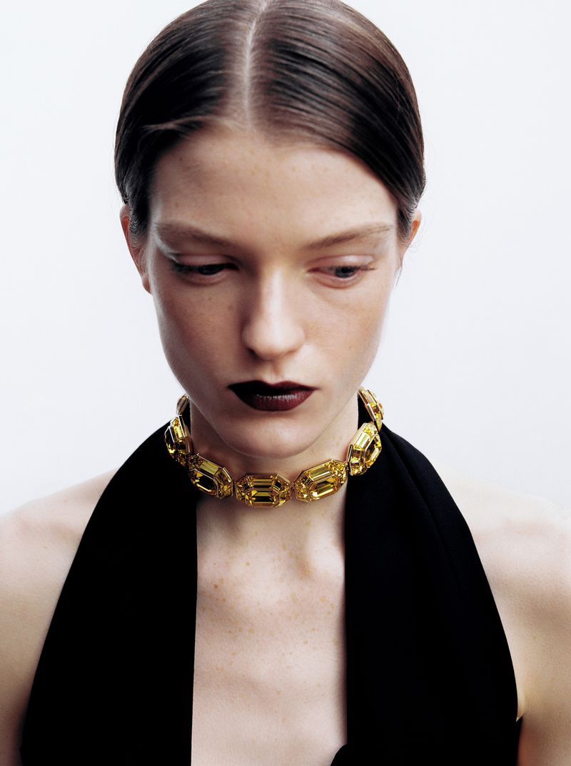 Close-up of a woman wearing a yellow stone choker, dark lipstick, and a black top against a white background in a beauty shot.