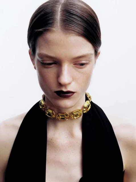 Close-up of a woman wearing a yellow stone choker, dark lipstick, and a black top against a white background in a beauty shot.