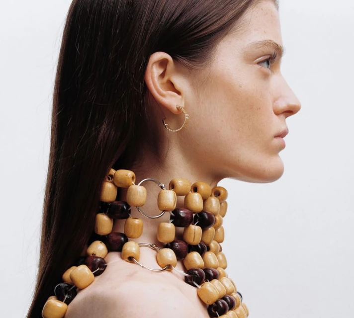 Profile shot of a light-skinned adult female with long brown hair wearing a wooden bead necklace against a plain white background.