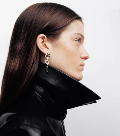 Profile portrait of a model with dark hair wearing a black leather jacket and dangling earrings against a white background.