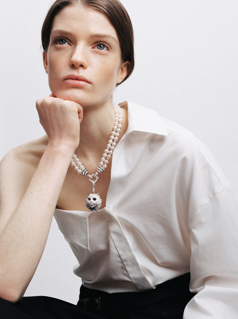 Model wearing a pearl necklace and white shirt against a plain light background