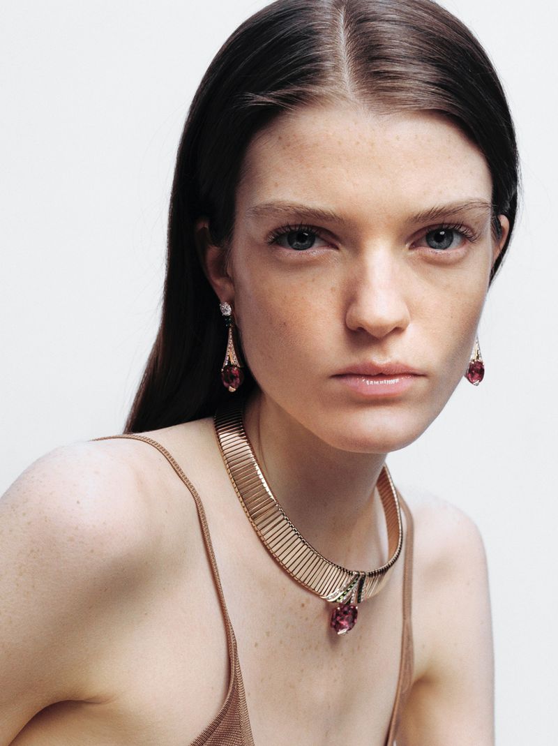 Close-up shot of an adult woman wearing a gold choker necklace and drop earrings with pink gemstones against a light background.