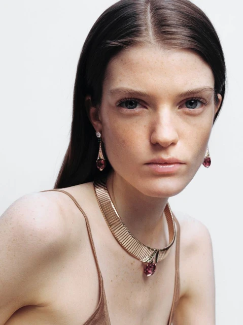 Close-up shot of an adult woman wearing a gold choker necklace and drop earrings with pink gemstones against a light background.
