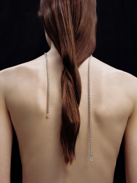 Back view of a slender person with long, braided brown hair wearing delicate silver necklaces against a dark studio backdrop.