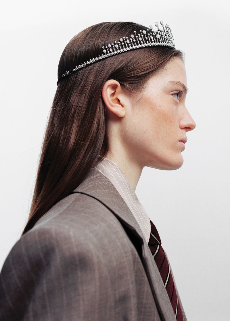 Woman in a pinstripe suit and tie, wearing a jewel-encrusted tiara, set against a plain white backdrop in a studio fashion setting.