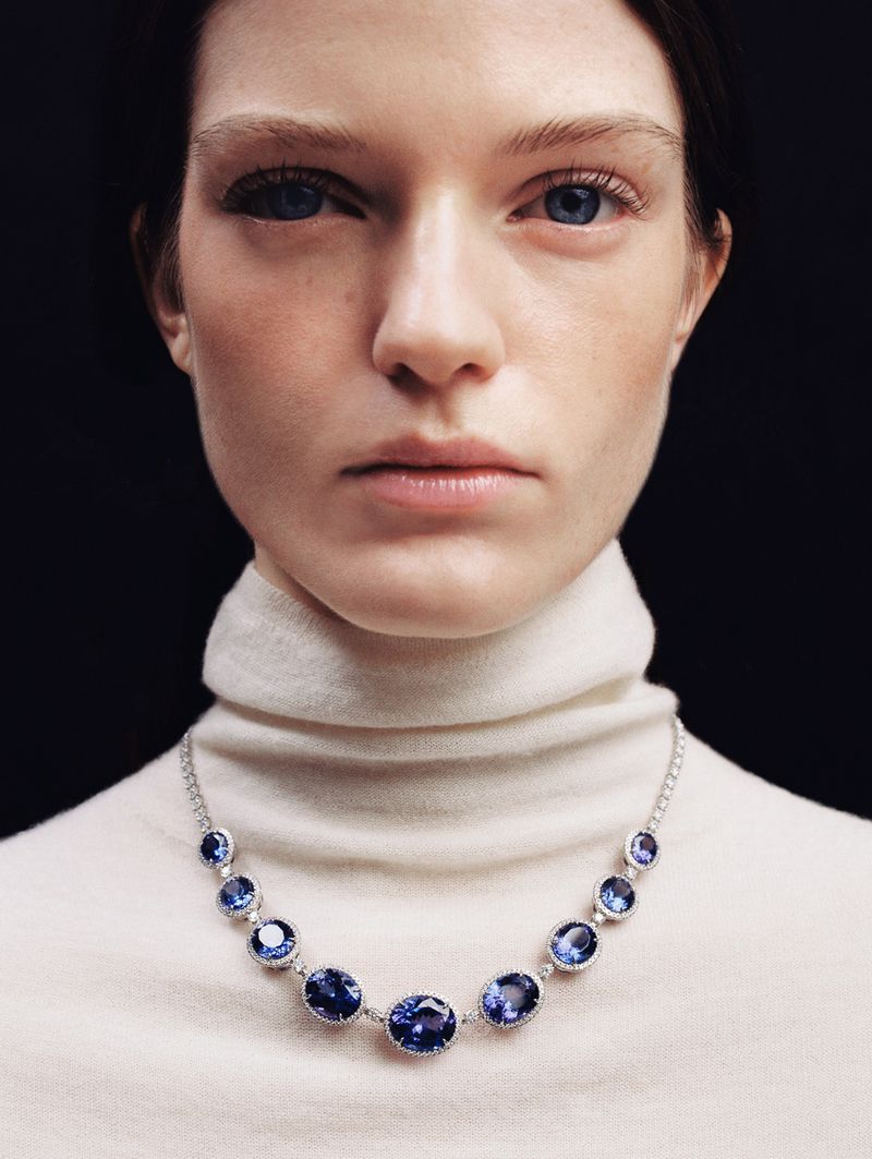 Close-up studio shot of a fair-skinned adult woman with blue eyes and a neutral expression, wearing a white turtleneck and a blue gemstone necklace.