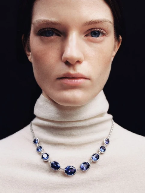 Close-up studio shot of a fair-skinned adult woman with blue eyes and a neutral expression, wearing a white turtleneck and a blue gemstone necklace.