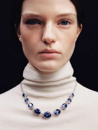 Close-up studio shot of a fair-skinned adult woman with blue eyes and a neutral expression, wearing a white turtleneck and a blue gemstone necklace.